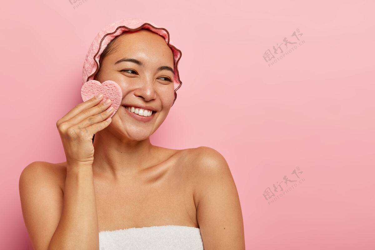 旁边容貌开朗的亚洲女人用化妆棉擦脸 卸妆 面容偏右 头戴防水保护帽 微笑温柔 牙齿洁白女士快乐光滑