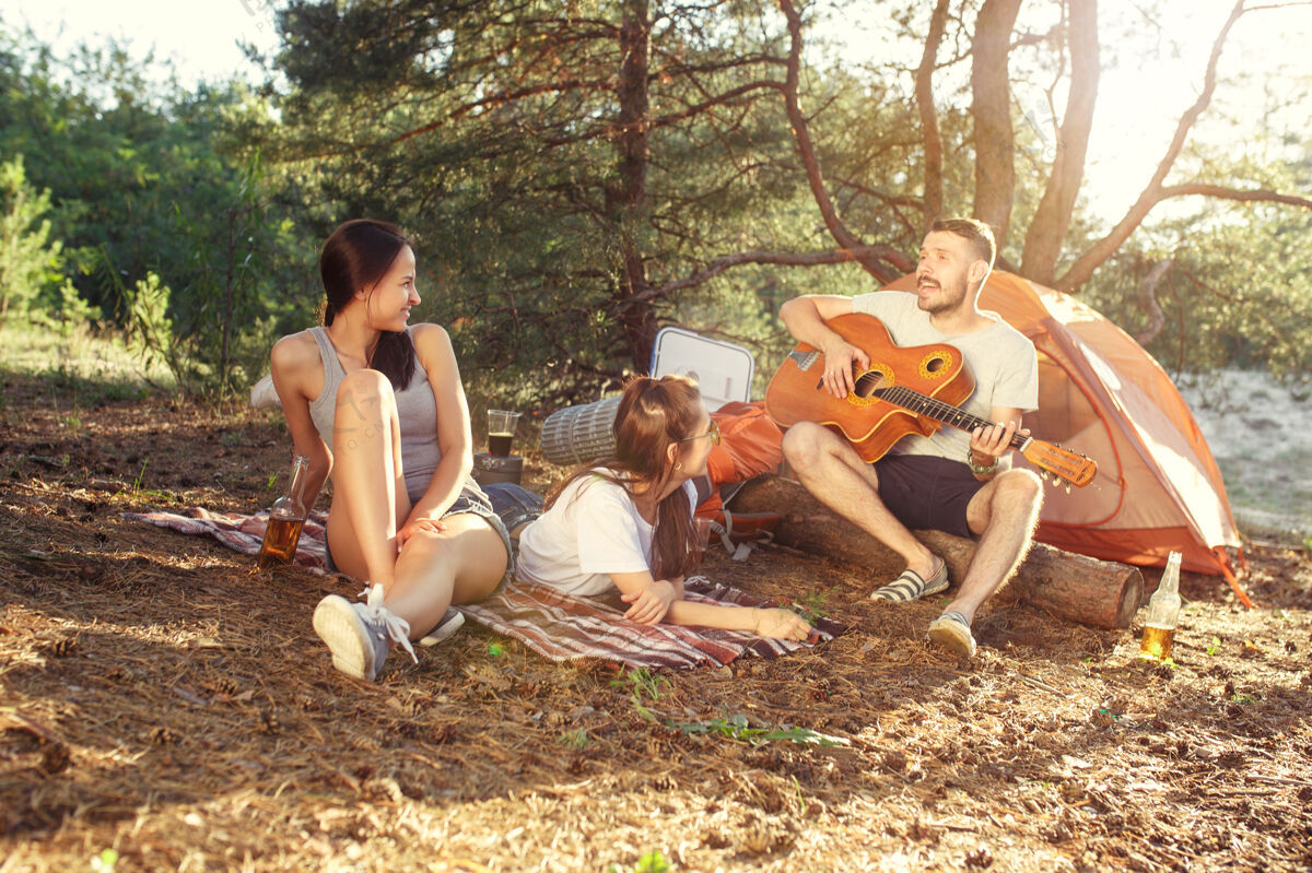 喝酒聚会 男女露营在森林里他们放松 唱着歌对抗绿草度假 夏天 冒险 生活方式 野餐的概念音乐乐趣团体