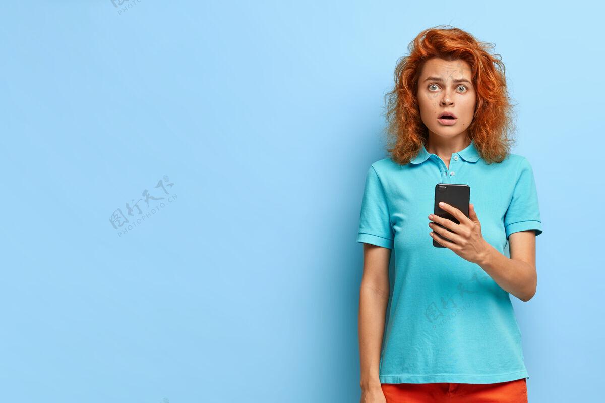 可怕惊艳美丽的欧洲女人 姜黄色的头发给人留下了深刻的印象 拿着现代手机 收到通知 穿着休闲服 模特们翻过蓝色的墙壁 留着复印空间欧洲红发室内