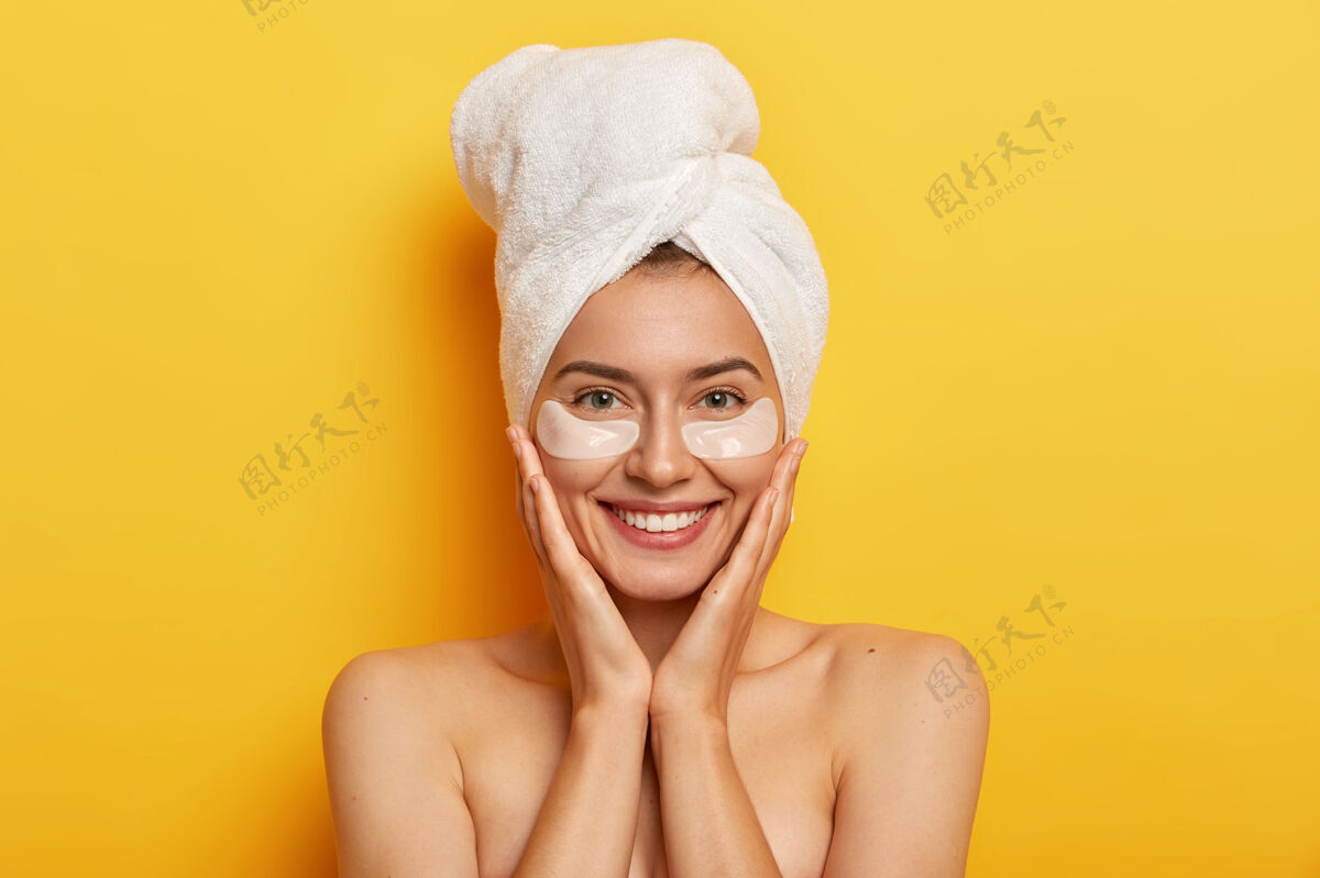 完美美丽开朗的女人用裹好的毛巾擦干头发 双手放在脸颊上 微笑温柔 皮肤清新 关心肤色 眼睛下面戴着眼罩情绪人高兴