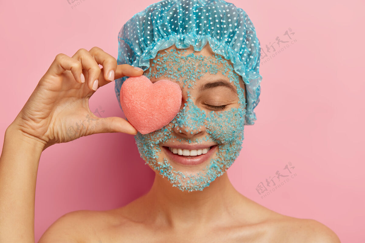 高兴开朗的年轻女子头上戴着水晶海盐磨砂膏 眼睛上拿着粉红色的心形海绵 积极地微笑 戴着淋浴帽 靠着粉色的墙壁做模特 从毛孔里剥脸盐室内满意