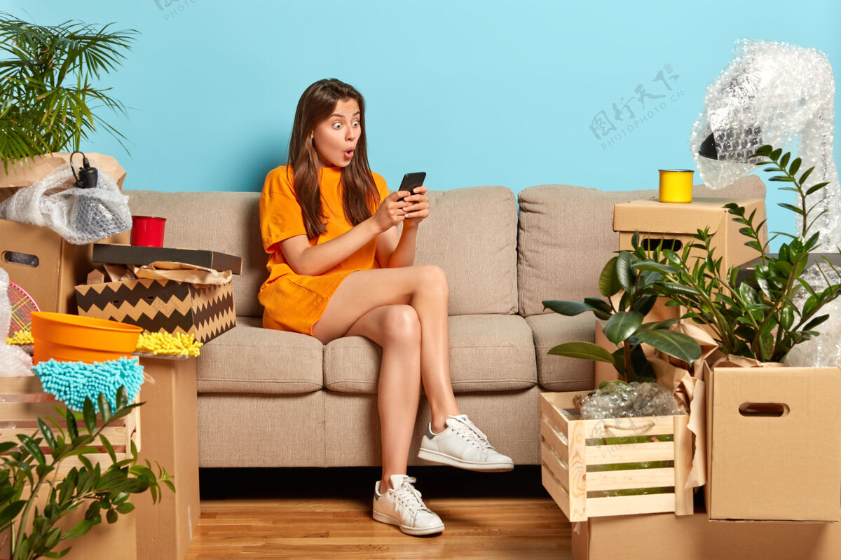 房主惊呆了的年轻白种人房主搬到新公寓 换了个家 盯着智能手机 计划改善家居 坐在舒适的沙发上 穿着橙色t恤 白色运动鞋印象单位昏迷