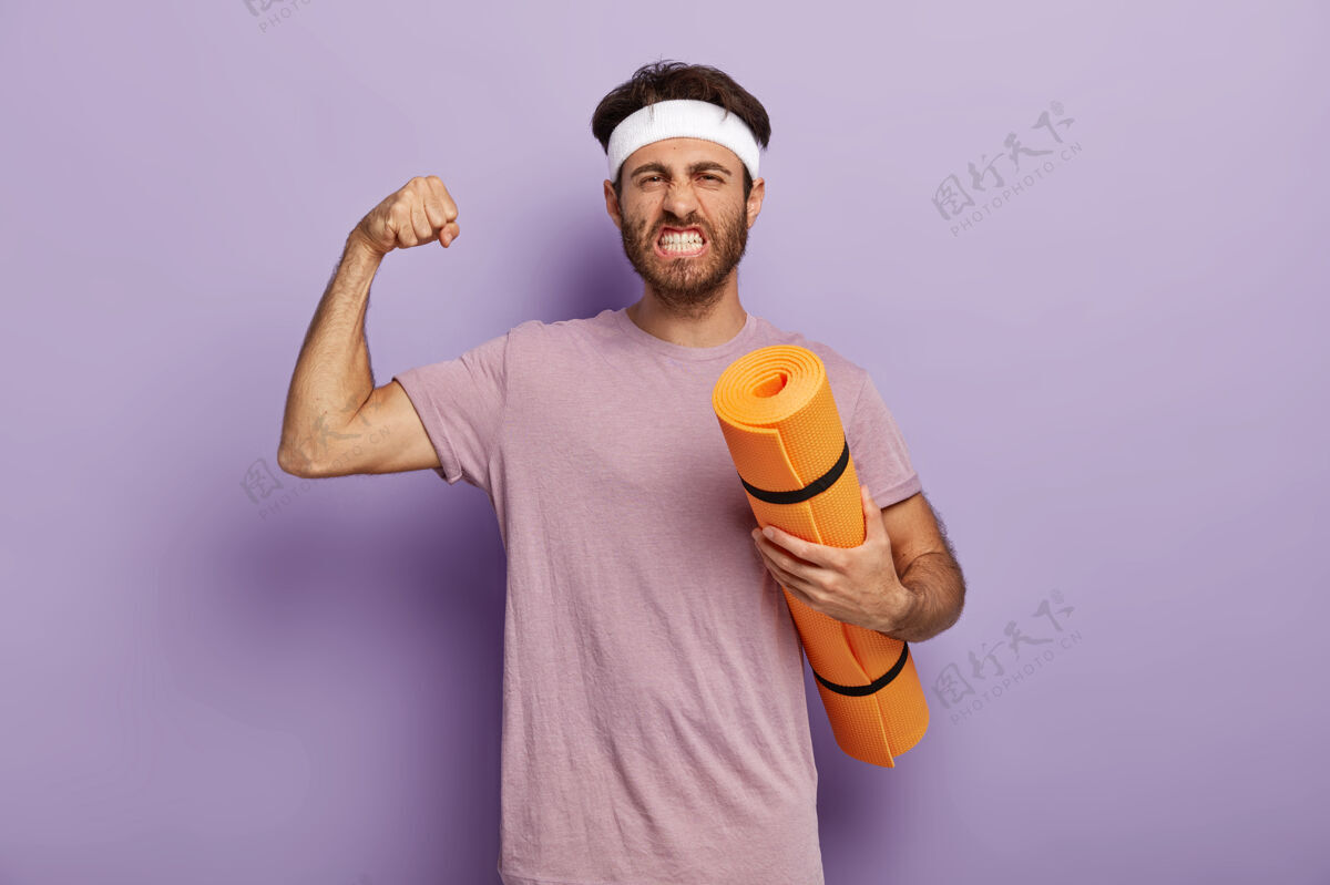 个人精力充沛的男人 拿着健身垫站着 把瑜伽作为运动和爱好来享受 举起手臂展示肌肉 咬紧牙关 戴着头带 穿着紫罗兰色t恤平衡你的生活 过健康的生活站着紫色锻炼