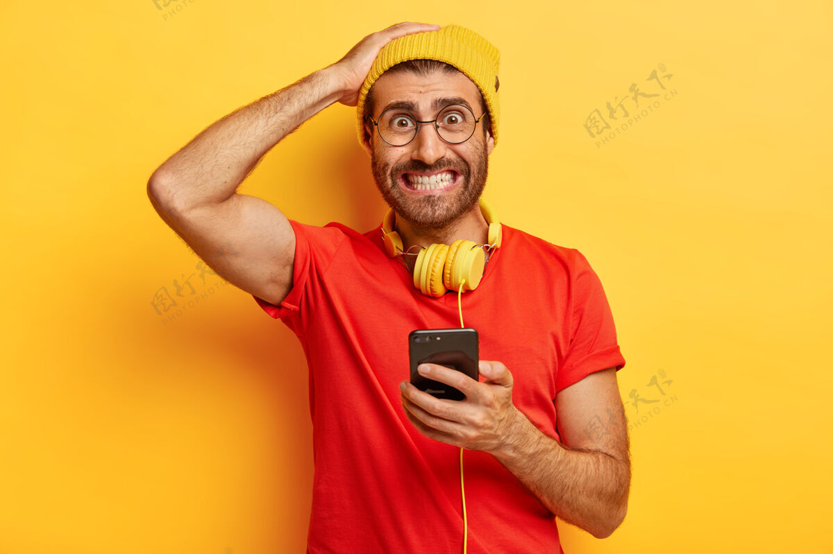 电话尴尬的潮人咬牙切齿 神情紧张 无法在智能手机上下载必要的应用程序 脖子上戴着耳机 穿着随意红色数字眼镜