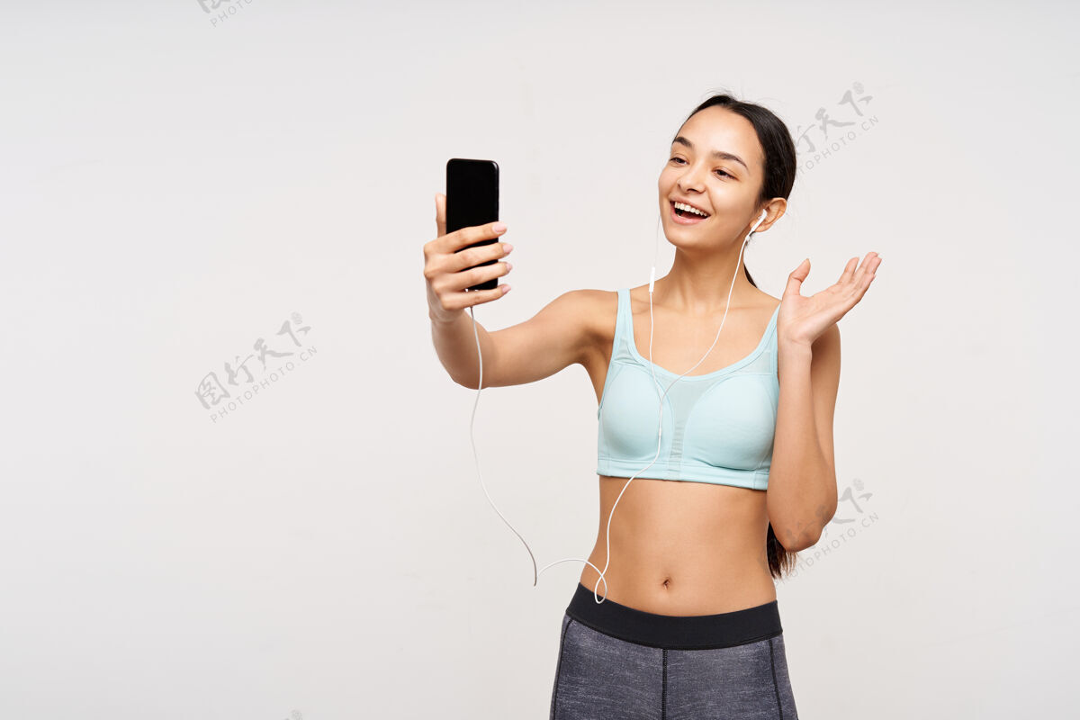 美容快乐的年轻运动棕色头发的女人微笑愉快 举手打招呼 同时与智能手机视频通话 隔着白墙孤立运动姿势头发