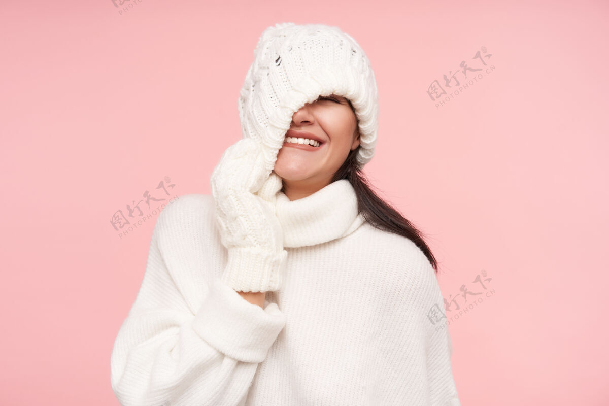 化妆品摄影棚拍摄的年轻快乐的黑发女人脱掉帽子 幸福地微笑着站在粉红色的墙上 穿着白色舒适的衣服高兴模特舒适