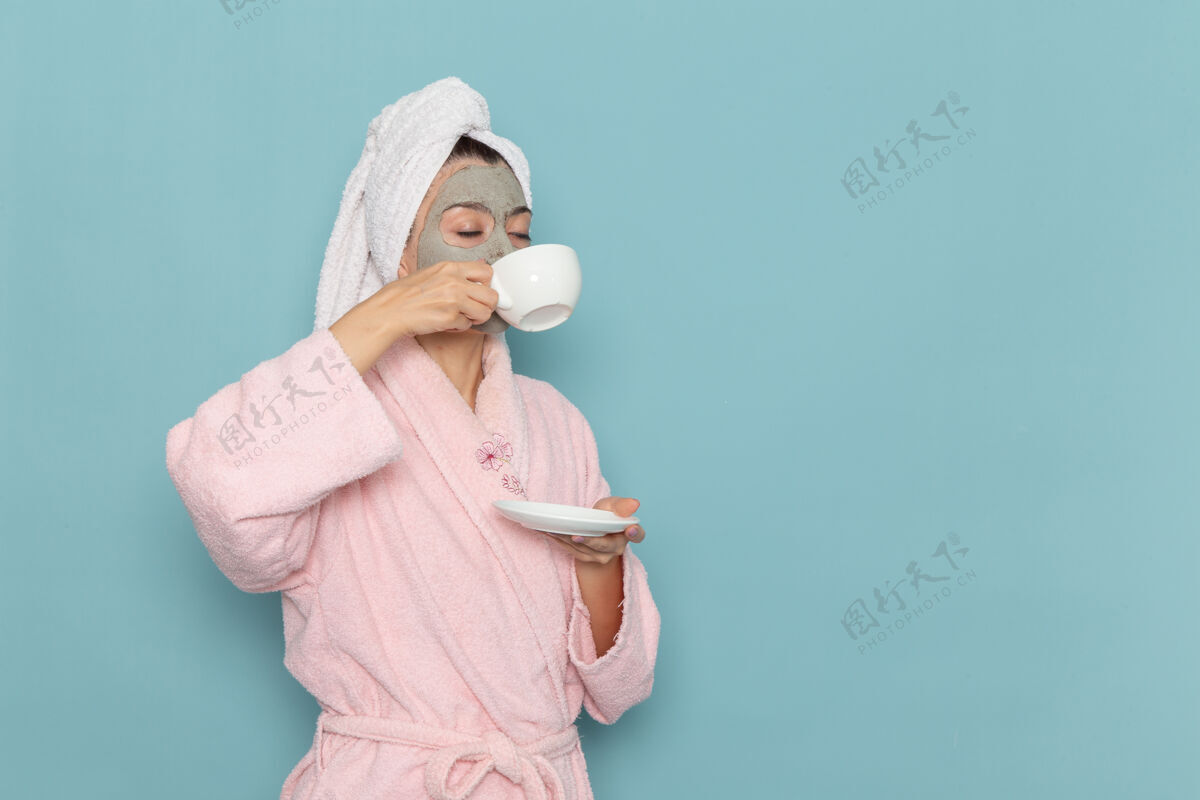 成人正面图穿着粉色浴袍的年轻女性在蓝色墙壁上喝咖啡清洁美丽干净的水自我护理霜淋浴女年轻饮用