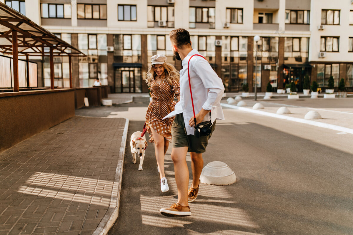 家庭穿着夏季服装的时尚夫妻在公寓的背景上和他们的狗一起奔跑和玩耍穿着浅色衬衫的男人牵着他心爱的手 拿着相机红领人圆点连衣裙