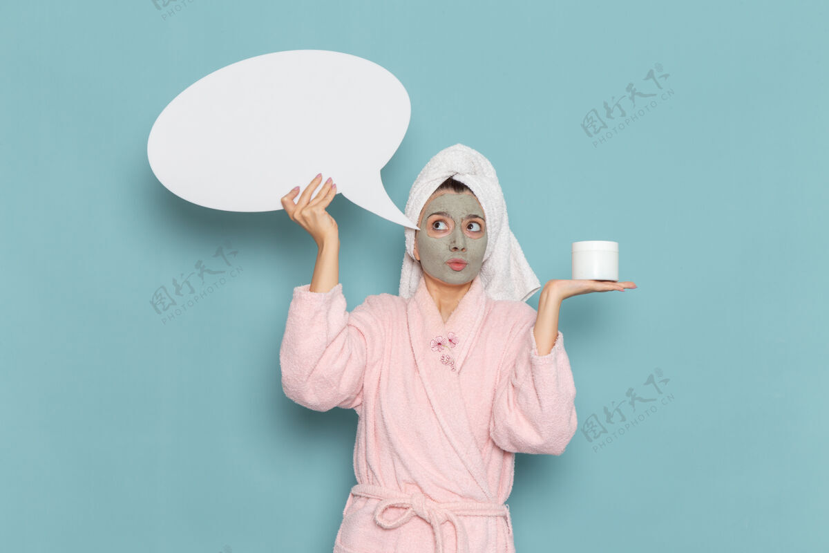 前面正面图身着粉色浴袍的年轻女性手持蓝色墙上巨大的白色标志淋浴清洁美容自理霜自我照顾清洁淋浴