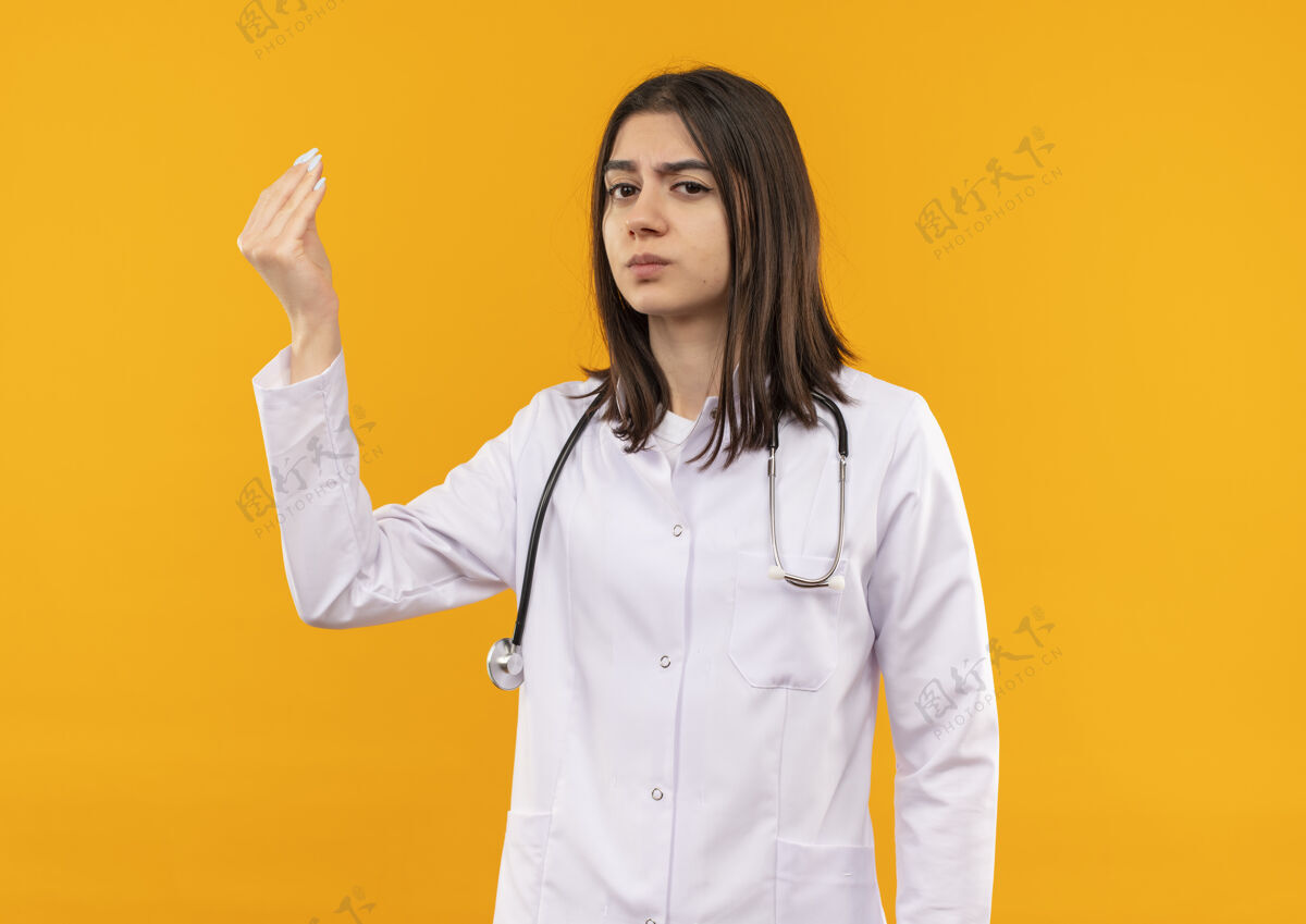 工作年轻的女医生穿着白大褂 脖子上戴着听诊器 站在橙色的墙上 不高兴地看着前面手医院女性
