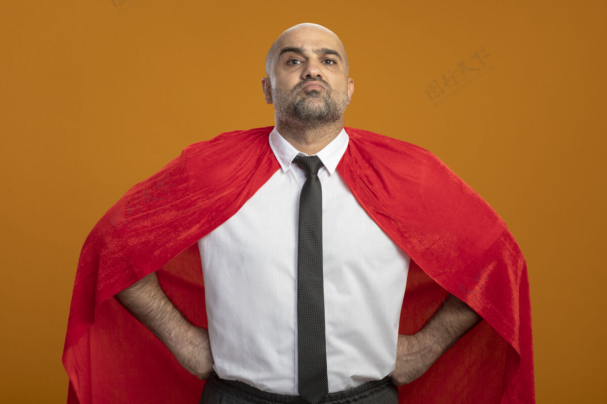臀部穿红色斗篷的超级英雄商人看起来很自信 双臂放在臀部站在橙色的墙上橙工作制服