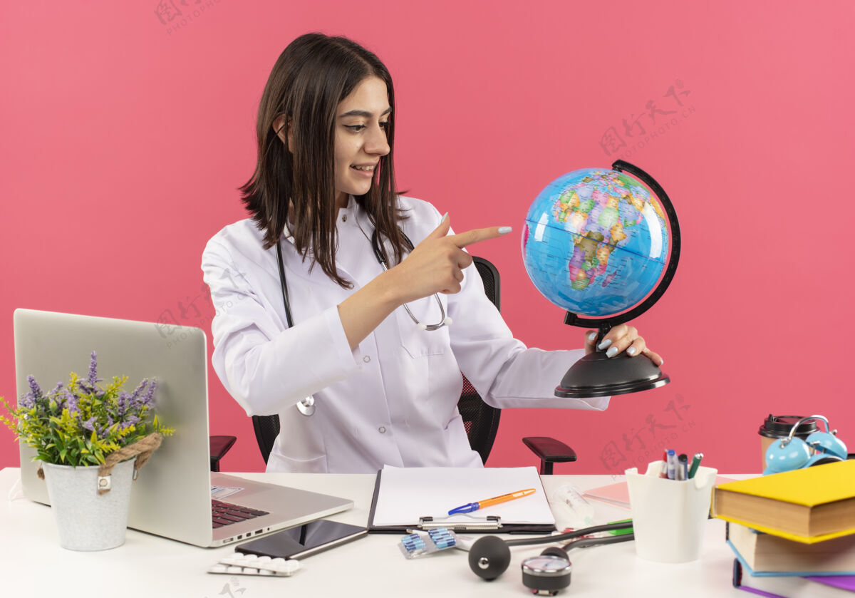脸年轻的女医生穿着白大褂 脖子上戴着听诊器 拿着地球仪 面带微笑地坐在桌旁 笔记本电脑放在粉红色的墙上笔记本电脑市民人