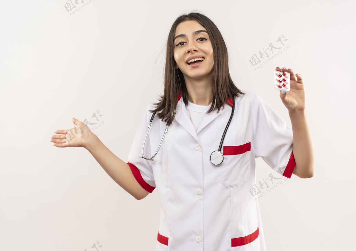 人年轻的女医生穿着白大褂 脖子上戴着听诊器 露出水泡 手里拿着药片指着旁边 站在白墙上又高兴又惊讶年轻手势惊喜