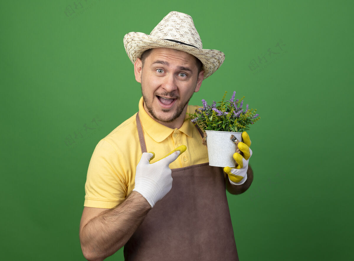 穿年轻的园丁穿着连体衣 戴着帽子 戴着工作手套 手里拿着盆栽植物 用食指指着它 站在绿色的墙边开心地笑着市民锅举行