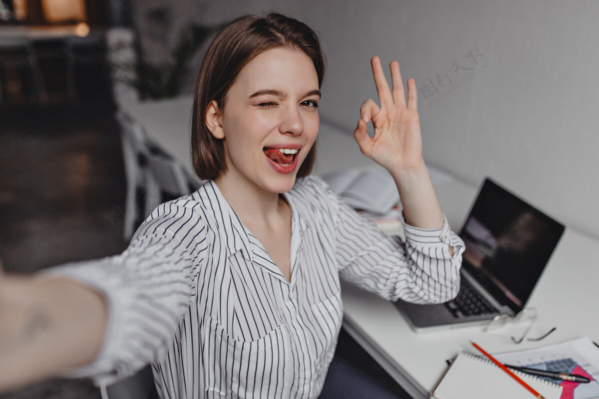 通信手上有纹身的淘气女人在办公室自拍穿着条纹衬衫的上班族在笔记本电脑的背景上显示“ok”标志女性微笑个人
