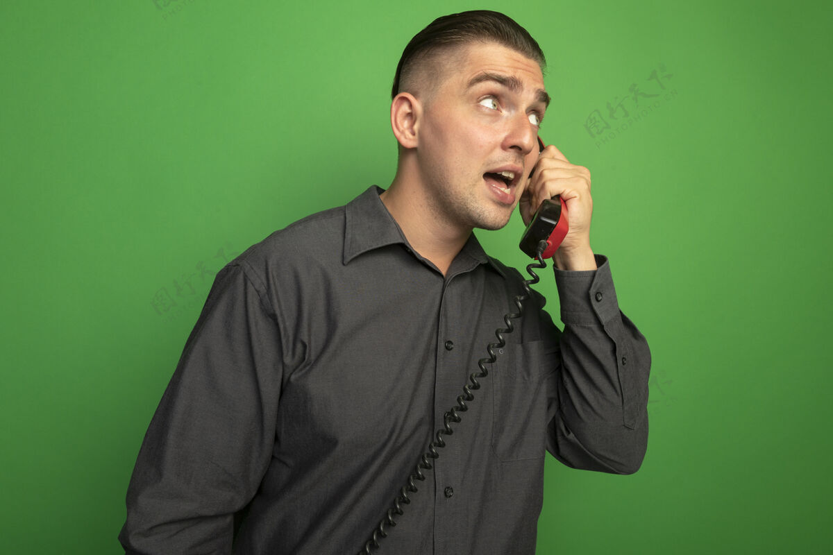 年轻穿着灰色衬衫的年轻帅哥正在讲老式电话 站在绿色的墙上困惑地看着一边人人看