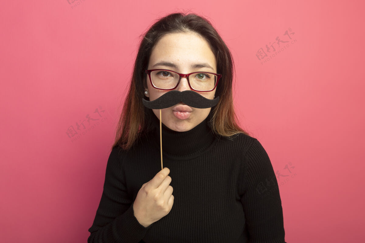 快乐一个穿着黑色高领毛衣 戴着眼镜的年轻漂亮女人站在粉红色的墙上 手里拿着一把滑稽的小胡子人高领毛衣棍子