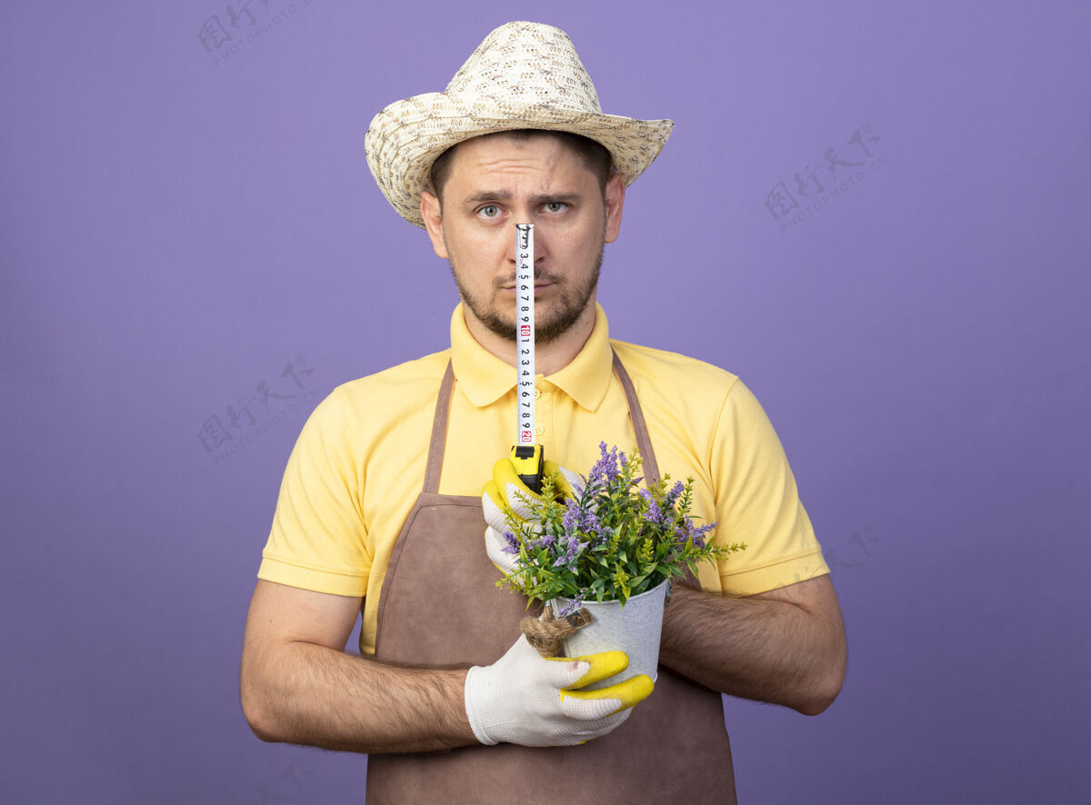 连身衣年轻的园丁穿着连体衣 戴着帽子 戴着工作手套 手里拿着盆栽植物和卷尺 严肃地站在紫色的墙上 看着前面锅紫色男人