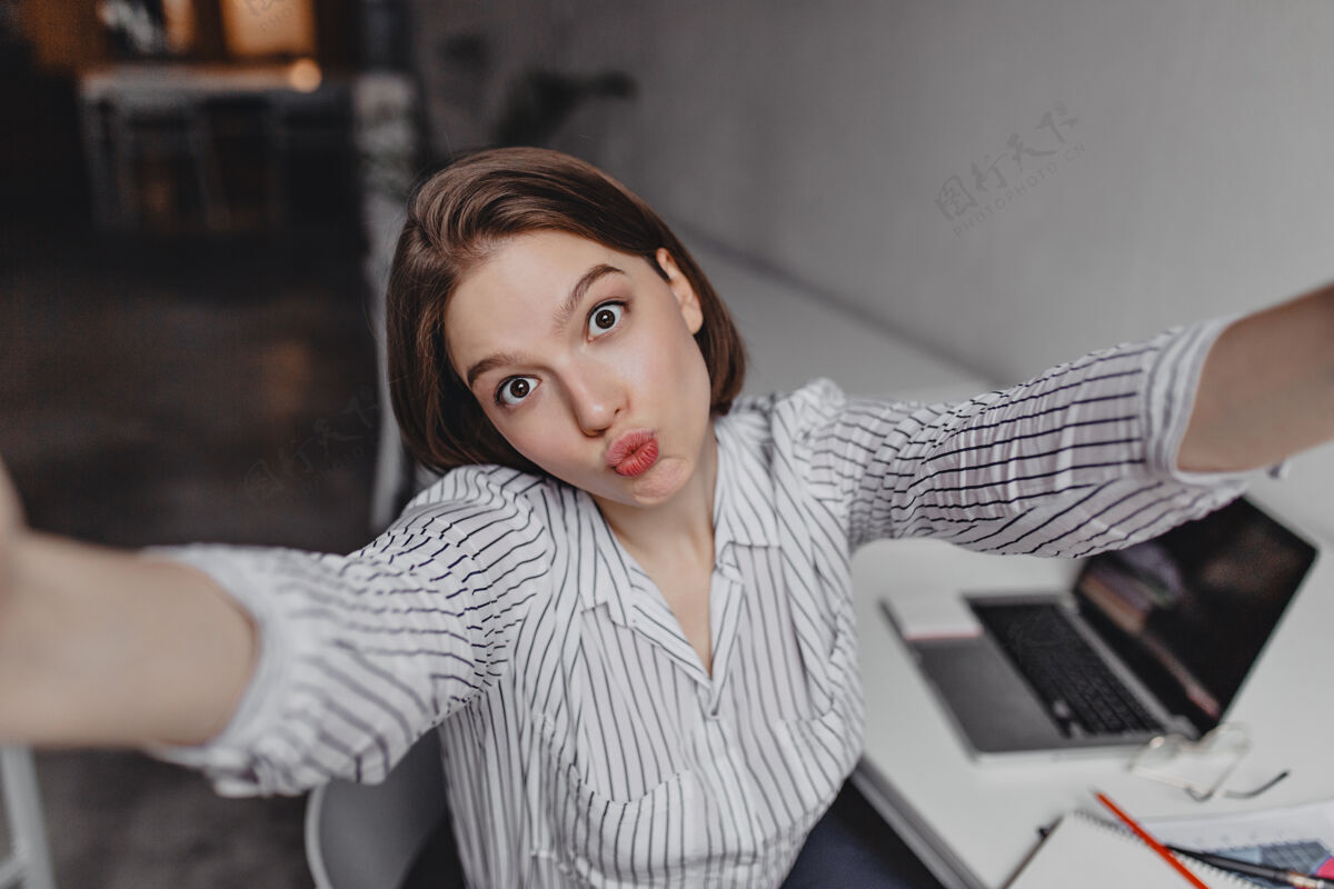 手机棕色眼睛的年轻女商人在白色的桌子和笔记本电脑上用滑稽的面部表情自拍电脑女孩办公室