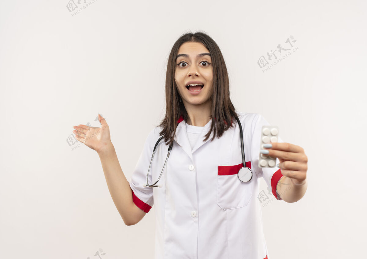 手势年轻的女医生穿着白大褂 脖子上戴着听诊器 露出水泡 手里拿着药片指着旁边 站在白墙上又高兴又惊讶医生女人年轻