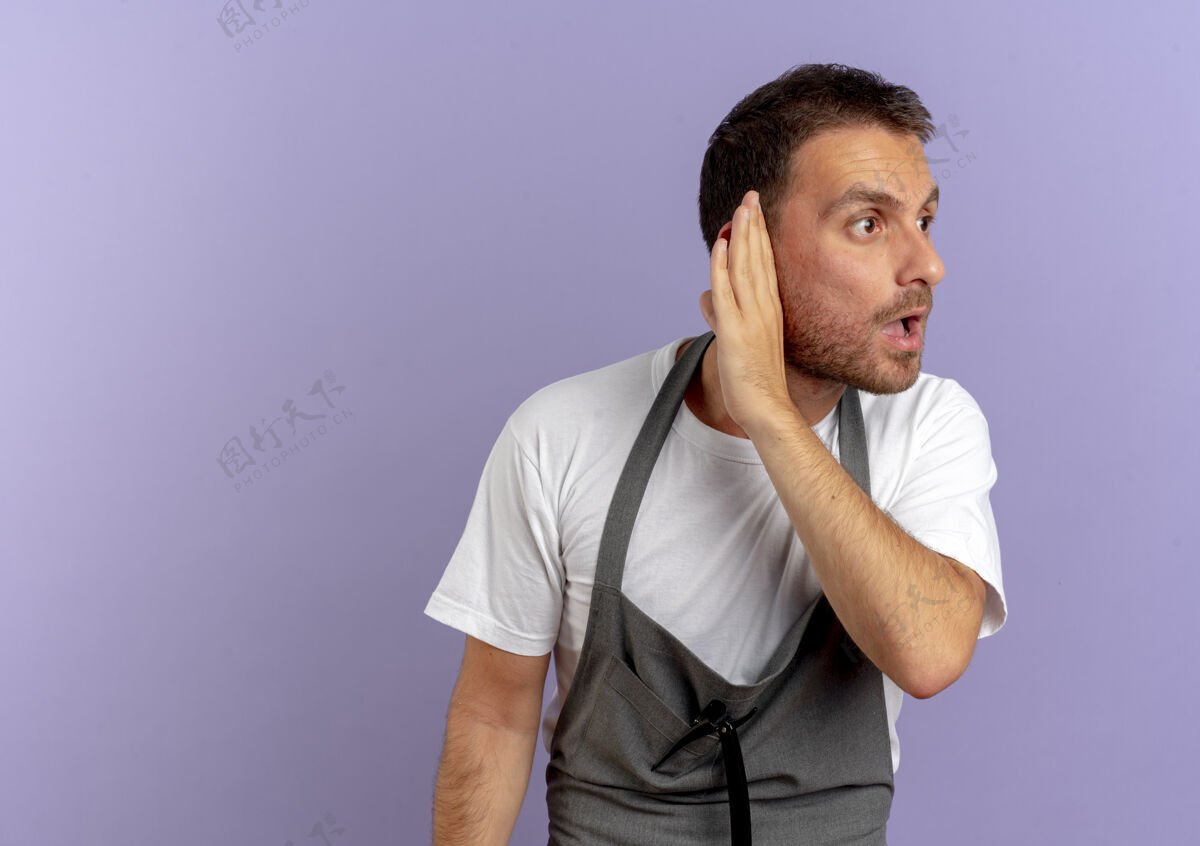 男人围裙上的理发师手里拿着理发机 手放在耳边 站在紫色的墙上试图倾听市民剪男人