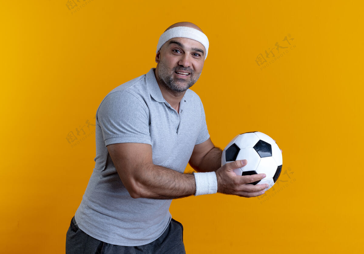 足球成熟的运动型男人戴着头巾 拿着足球 面带微笑地站在橙色的墙上望着前方抱着微笑站着