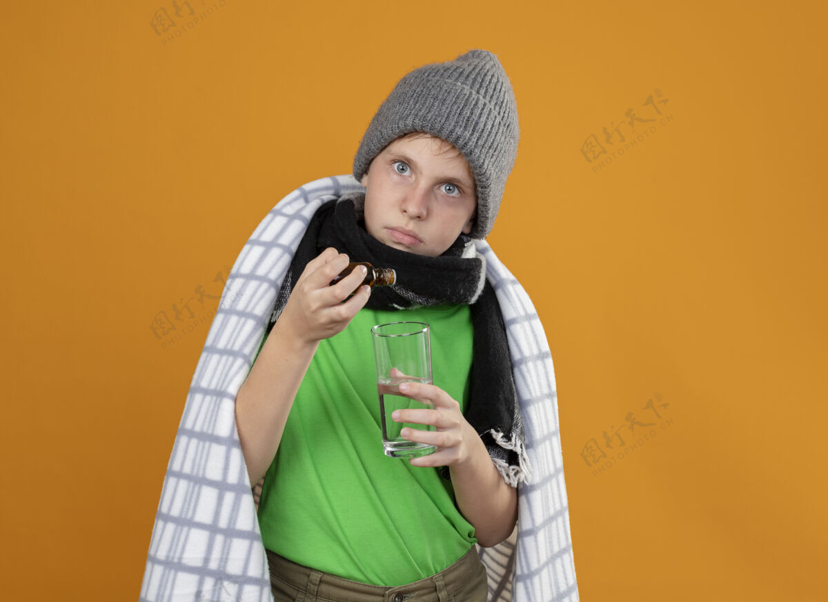 滴生病的小男孩戴着暖和的帽子 围着围巾 裹在毯子里 从药瓶里滴下来的药水滴在玻璃上 站在橘黄色的墙上玻璃橘子帽子