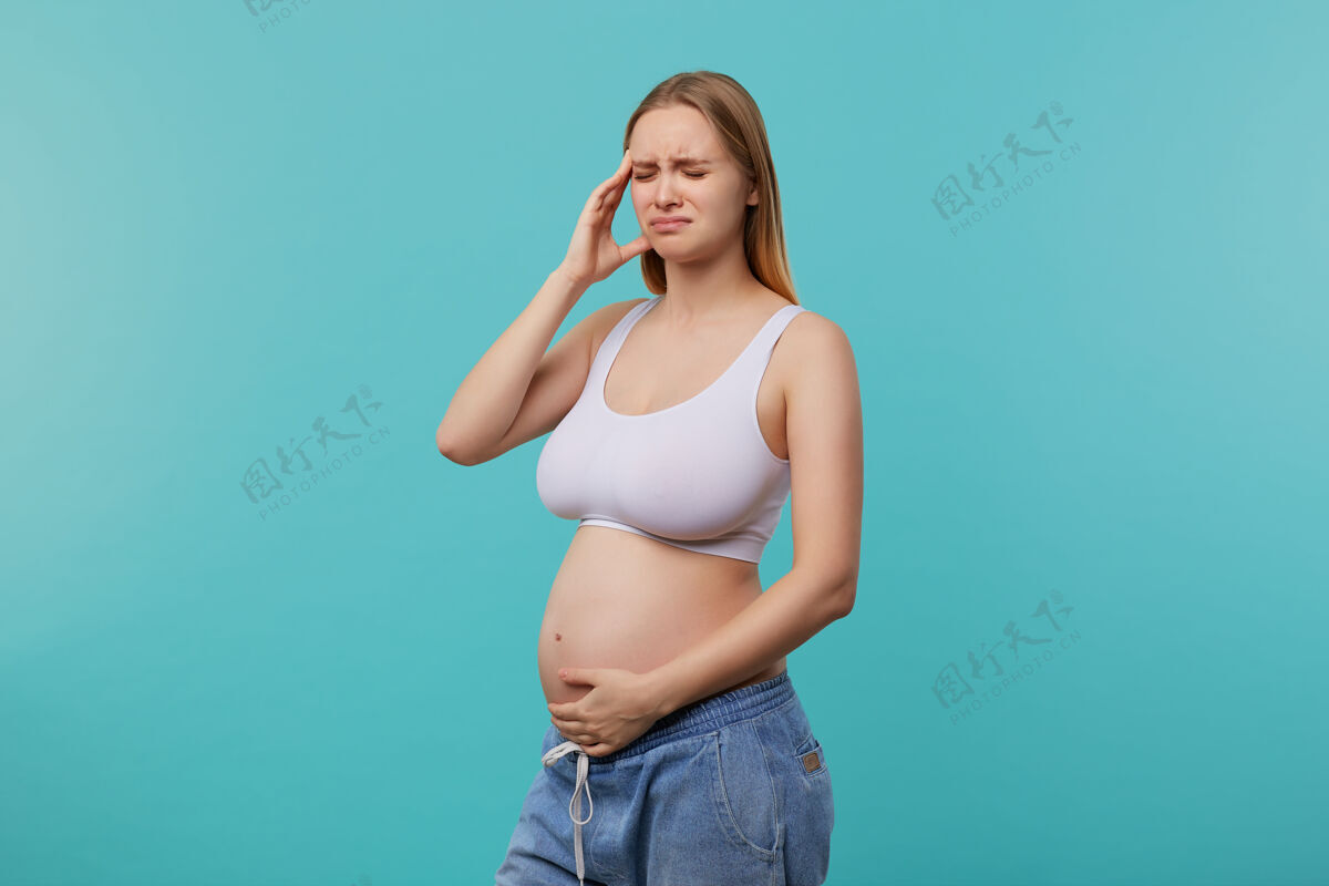 站压力重重的年轻金发长发未来的妈妈举起手放在肚子上 闭着眼睛愁眉苦脸 站在蓝色的背景下情绪女性女士