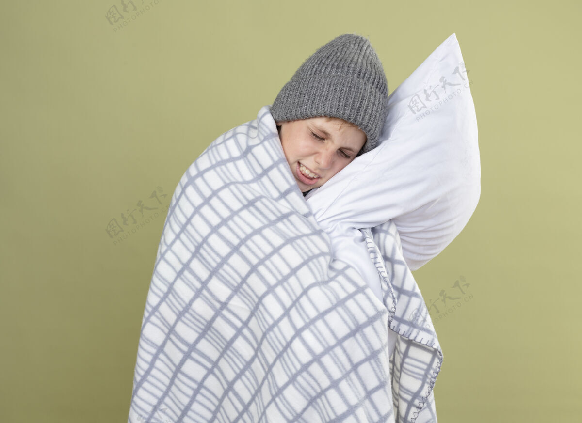帽子生病的小男孩 戴着暖和的帽子 裹着毯子 抱着枕头 站在轻墙上感觉不舒服光站着枕头