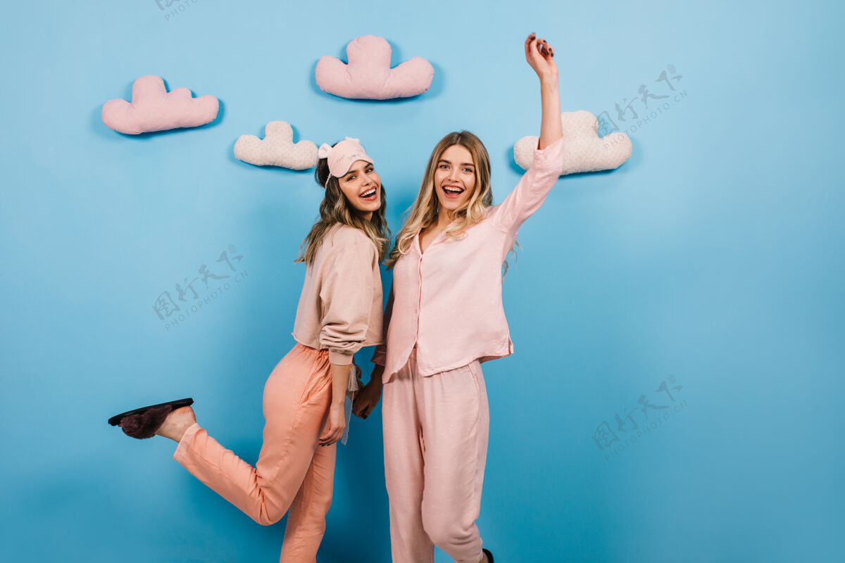 年轻穿着睡衣的兴奋姐妹们在蓝色的墙上和玩具云跳舞友谊姐妹兴奋