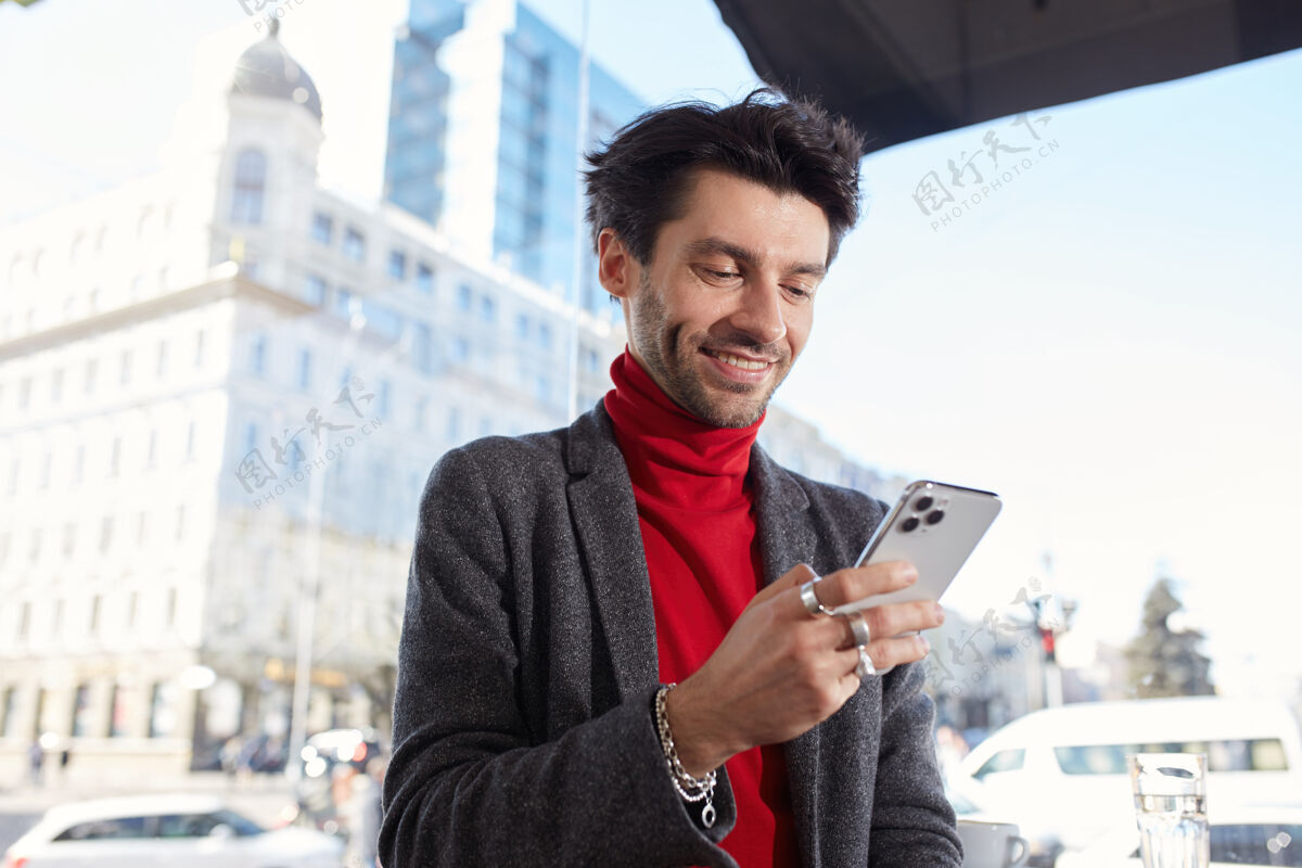 优雅站在城市的背景下 快乐的年轻优雅的黑发胡须的家伙拿着手机 高兴地微笑着查看他的社交网络正面短裤咖啡馆