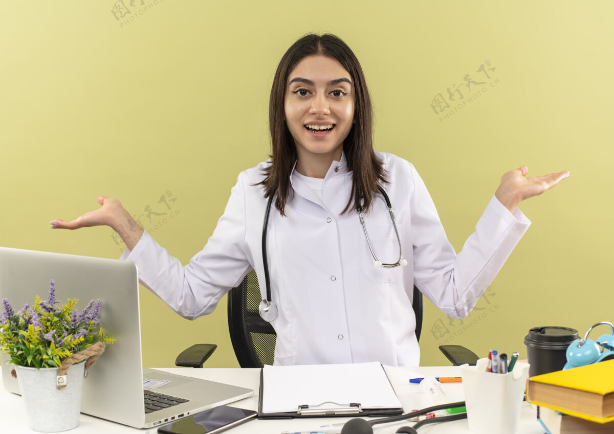 桌子年轻的女医生穿着白大褂 脖子上戴着听诊器 面带微笑地看着前面 双臂伸向两边 坐在桌子旁 笔记本电脑放在浅色的墙上人人坐着