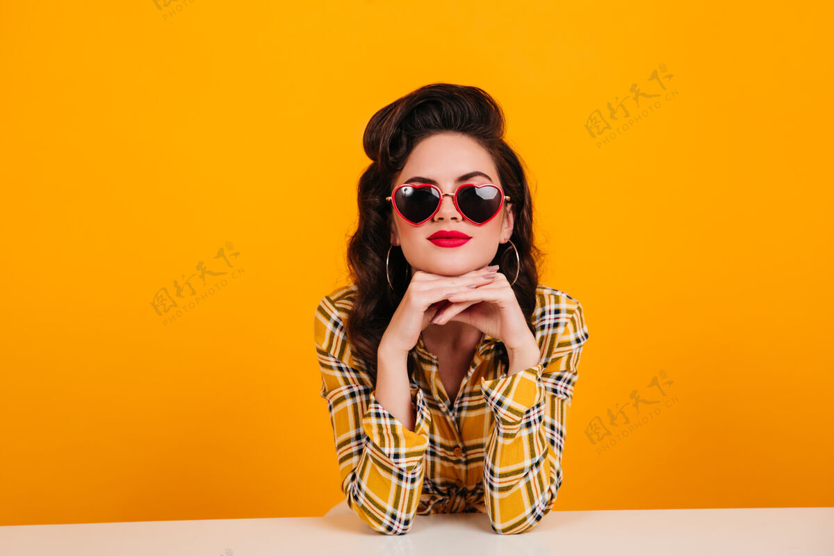 深色好奇的年轻女子坐在黄色背景上摄影棚拍摄的戴着心形眼镜的漂亮小姑娘优雅成人微笑