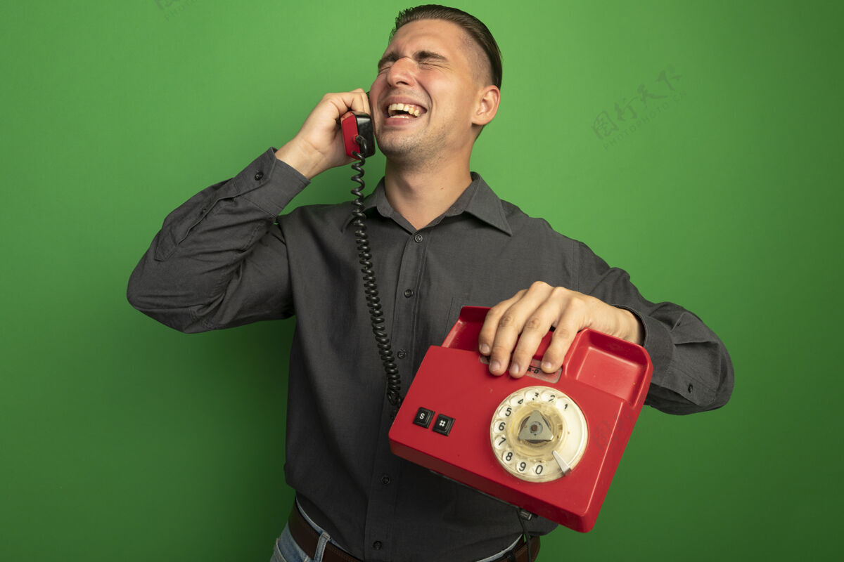人穿着灰色衬衫的年轻帅哥站在绿色的墙壁上 兴高采烈地讲着老式电话电话欢呼英俊