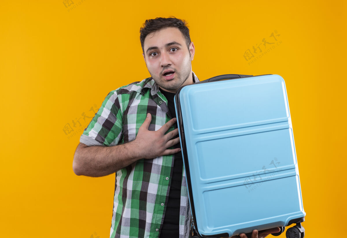 旅行站在橙色的墙上 一个穿着格子衬衫的年轻人拿着手提箱 看上去既惊讶又困惑站着看抱着