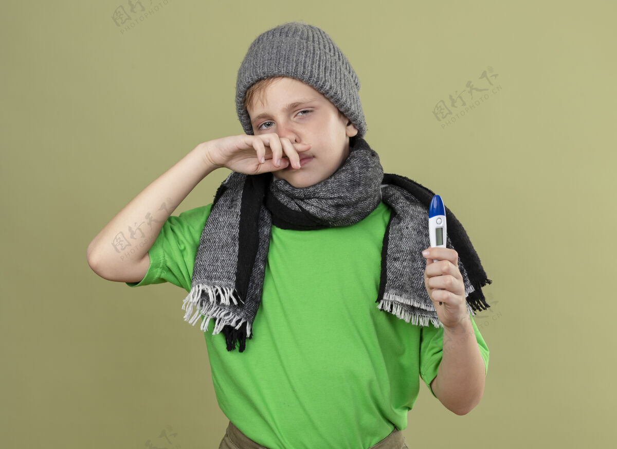 光生病的小男孩穿着绿色t恤 戴着暖和的围巾和帽子 感觉不舒服拿着温度计擦鼻子站在光墙上流鼻涕体温计帽子擦