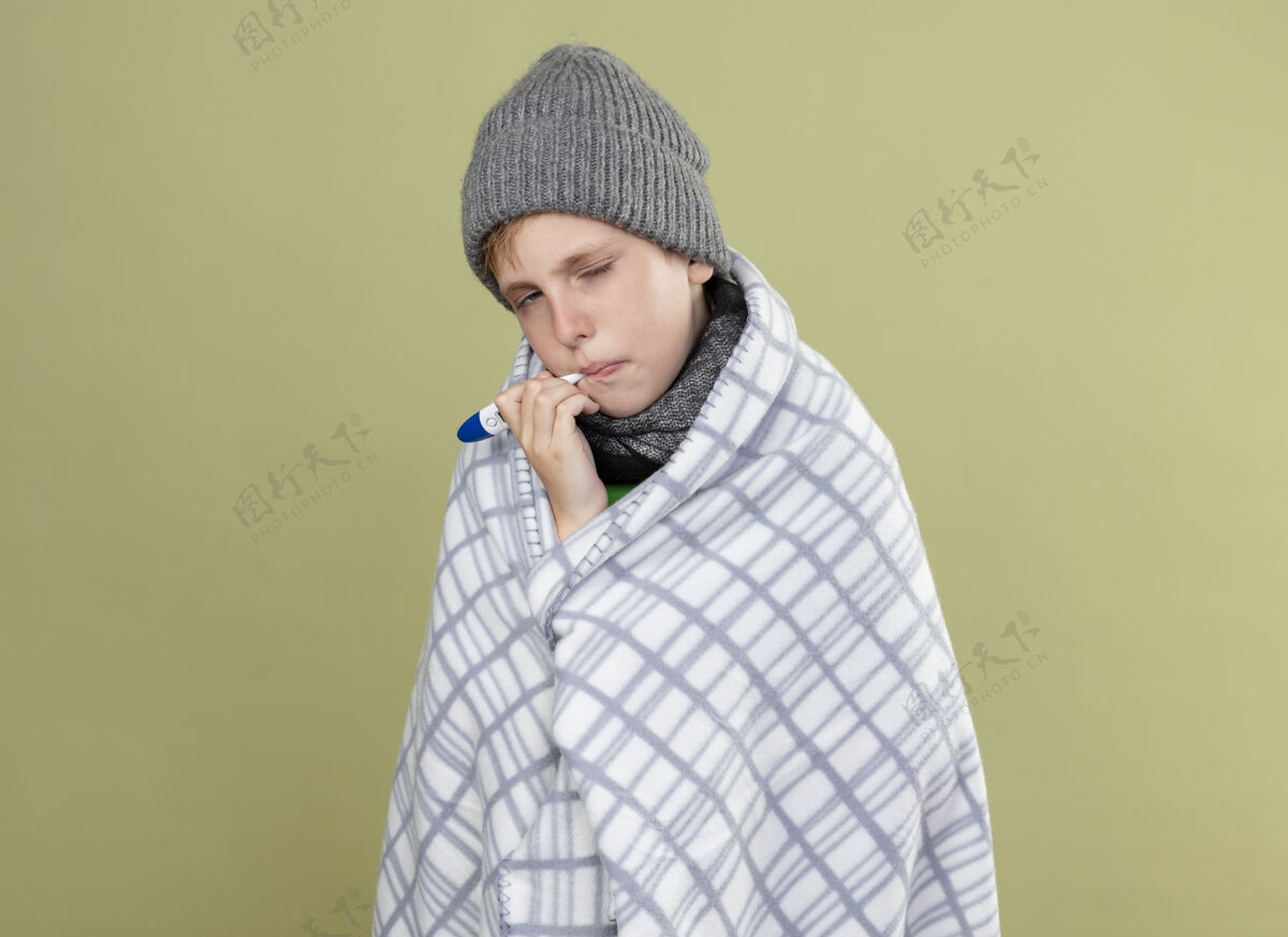 测量生病的小男孩穿着绿色t恤 戴着暖和的围巾 帽子裹着毯子 把温度计放进嘴里 站在光墙上测量温度温度计支架灯