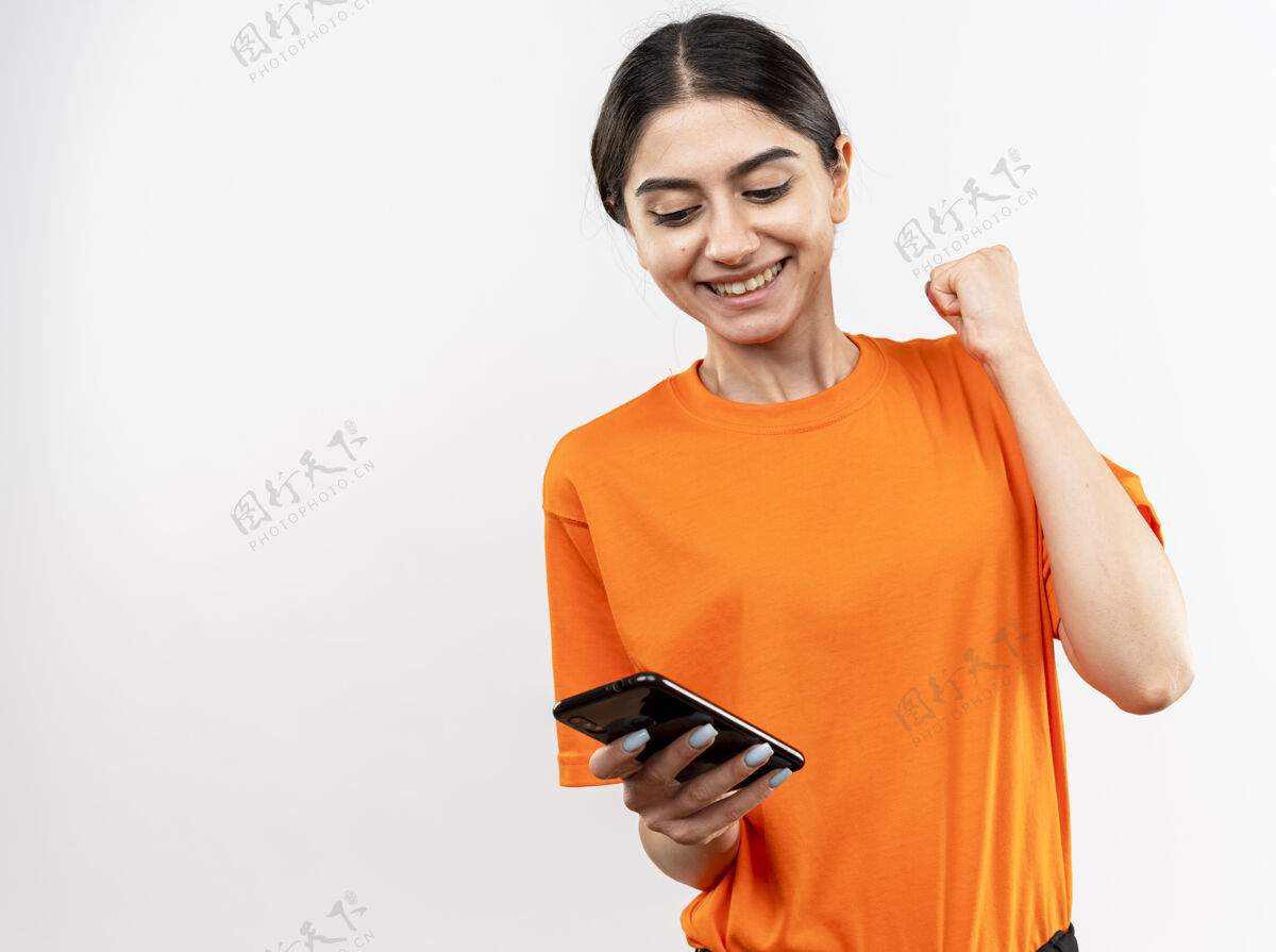 欢呼身穿橙色t恤的年轻女孩手持智能手机握紧拳头 高兴而兴奋地微笑着 为自己的成功站在白墙上而欢欣鼓舞穿年轻高兴