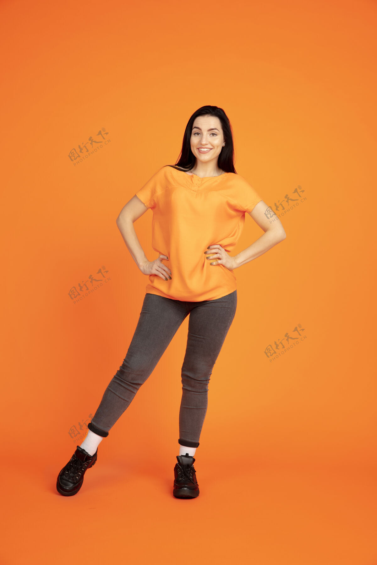 专业橙色工作室背景上的白种人年轻女子肖像穿着衬衫的漂亮黑发女性模特人类情感的概念 面部表情 销售 广告广告空间站立和微笑英俊工作公司