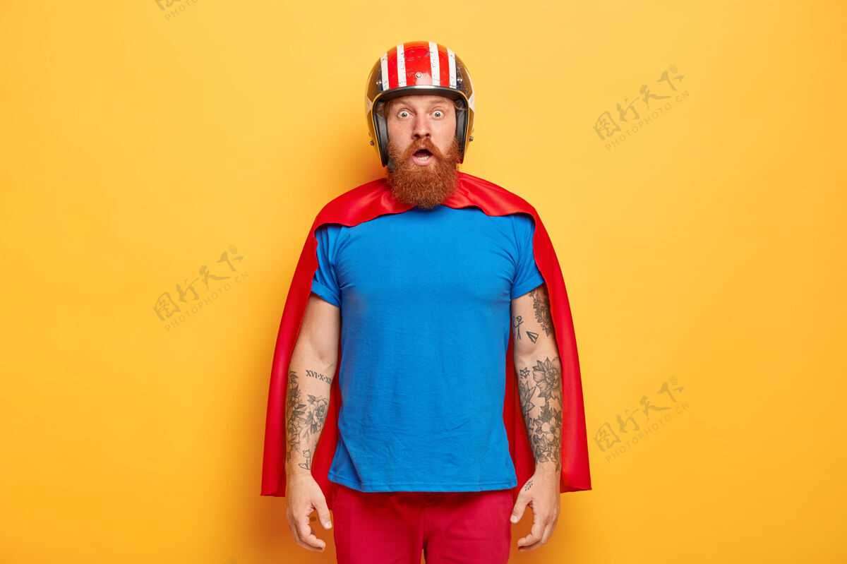 姜情绪化的姜胡子男人戴着防护头盔 红色斗篷和蓝色t恤害怕斗篷飞行