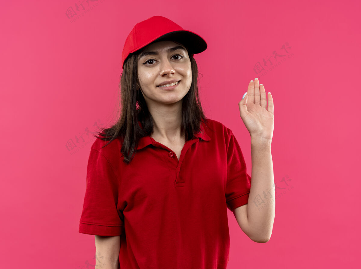 站着穿着红色马球衫 戴着帽子的年轻送货女孩微笑着 幸福地站在粉红色的墙上挥手致意马球挥手微笑