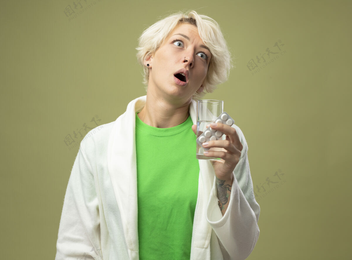 抱着一个短发的生病女人站在明亮的墙壁上 手里拿着一杯水 手里拿着一个水泡 手里拿着药片 看着旁边女人疾病感觉