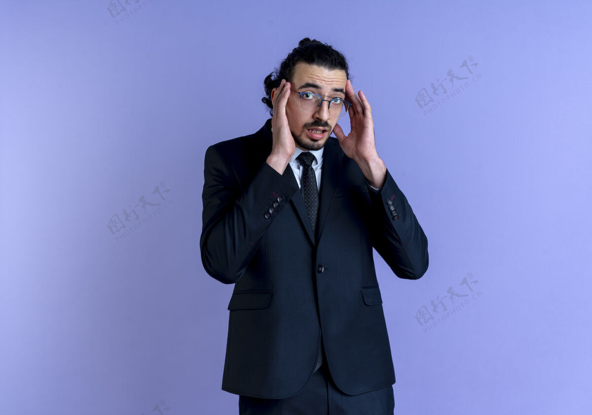 生意人一个穿着黑西装戴着眼镜的商人望着前面 担心地抚摸着站在蓝色墙壁上的太阳穴站西装男人