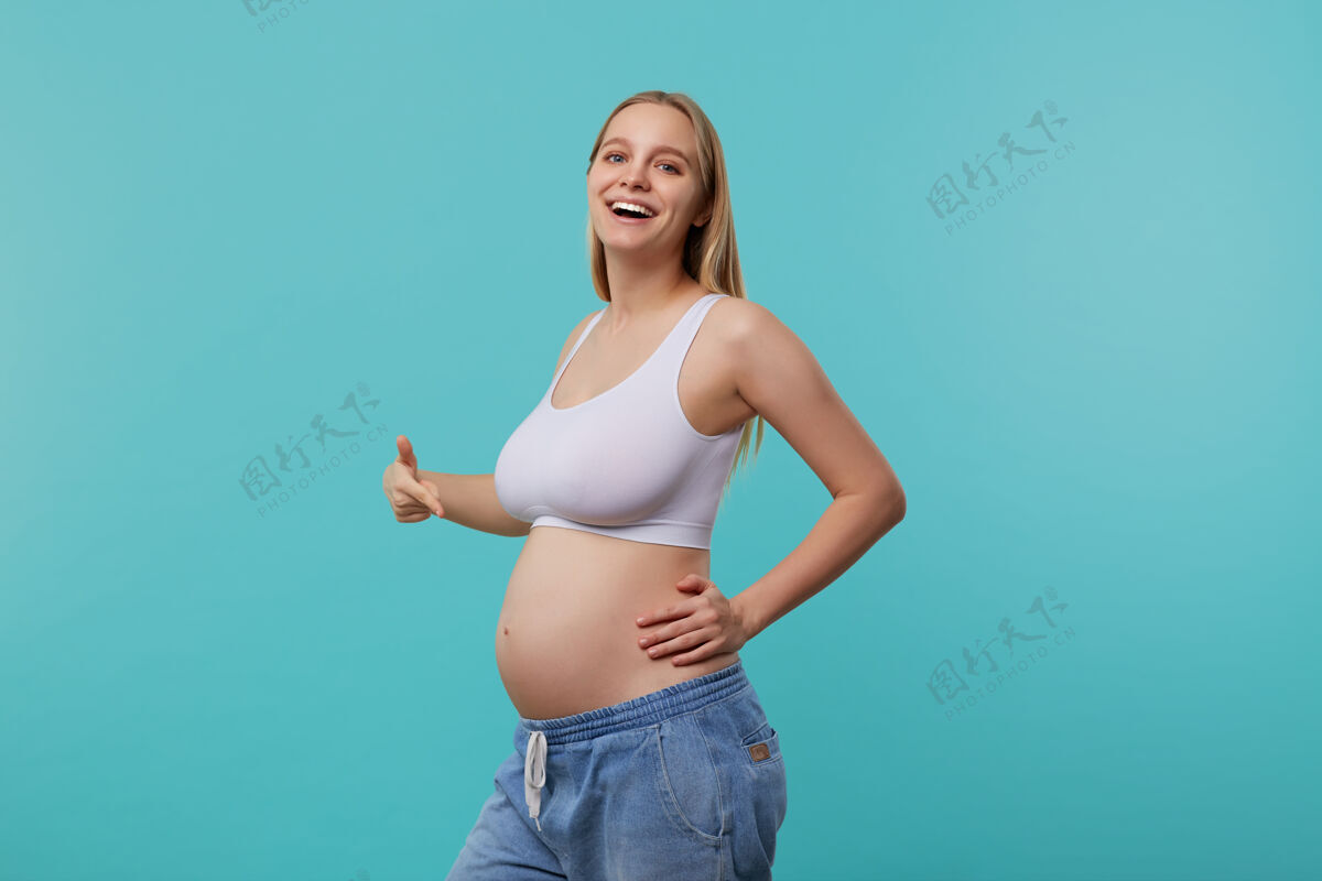 上衣室内照片 年轻可爱的金发孕妇 自然妆容 在她的肚子上愉快地举起手 微笑广泛 孤立在蓝色背景发型妈妈衣服