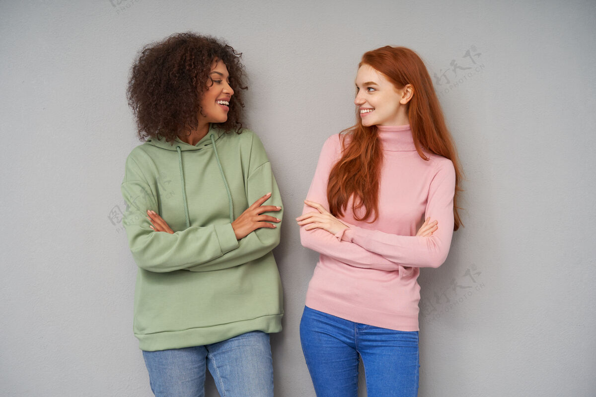 微笑迷人欢快的年轻女士们面带微笑 面带微笑 双手交叉放在胸前 在灰色的墙壁上摆姿势衣服模特编织