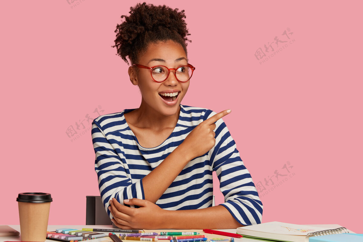 内容艺术文具表情开朗的快乐黑人妇女 戴眼镜以获得良好的视觉效果 指着右上角 注意到一些惊人的东西 用笔记本 彩色铅笔作画眼镜写作满意
