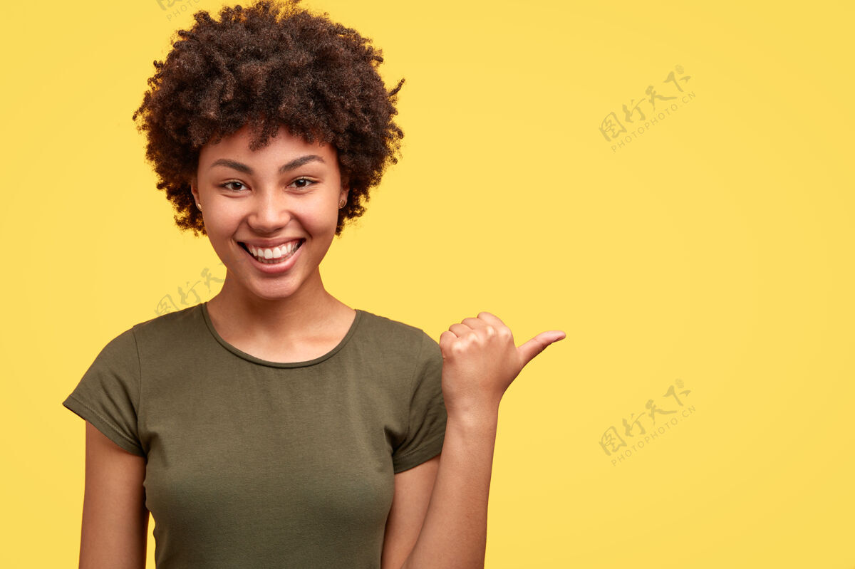 青少年好笑的美籍黑人女性正面朝右点 穿着休闲t恤 在黄色墙壁上展示自由空间 建议去那里 很友好手指内容高兴