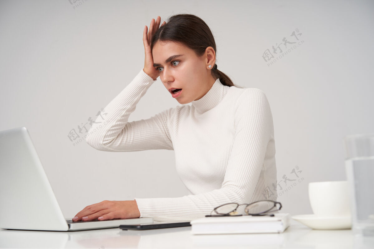 惊人压力重重的年轻黑发美女 穿着白色针织马球衫 在白墙上拿着笔记本电脑 一脸震惊地读着意想不到的新闻坐着电话女士