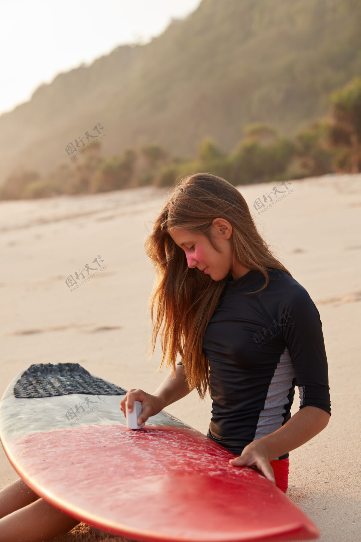 沙滩户外拍摄的年轻女孩在冲浪板上打蜡以确保安全乘浪 穿着黑色泳衣 坐在温暖的沙滩上 注意安全 眼睛周围涂上粉色锌 享受自由休闲概念高加索活跃天空
