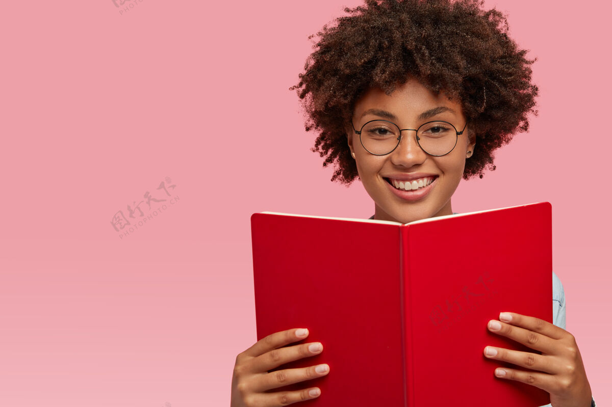 快乐快乐快乐的非洲裔美国妇女 表情愉快 戴着视力好的眼镜 拿着课本 为研讨会学习材料 隔着粉红色的墙壁 左边是空白微笑教育文学